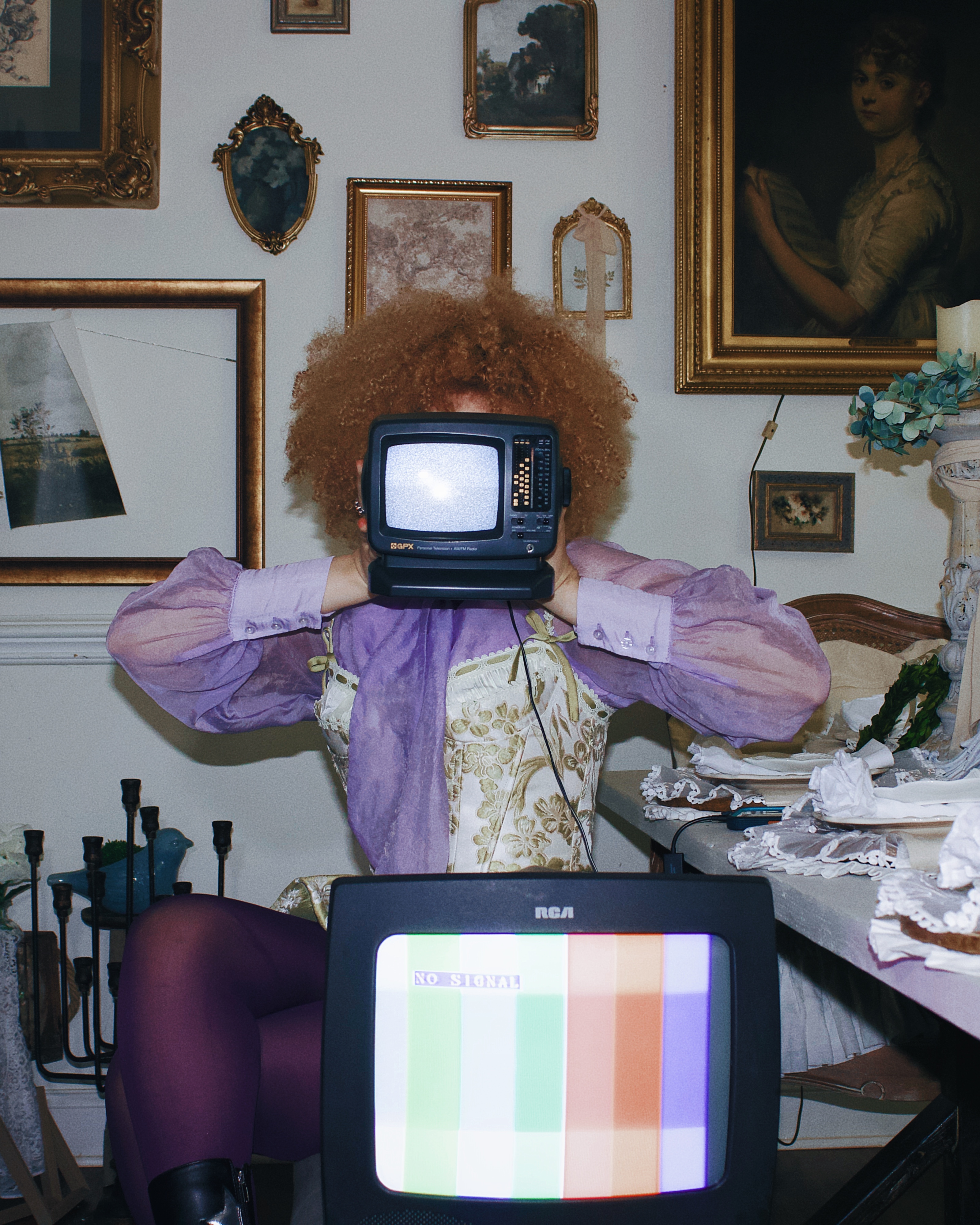 A person with curly hair holds a vintage TV in front of their face, sitting in a room decorated with framed art on the walls. A second TV displays a 'No Signal' screen in the foreground.