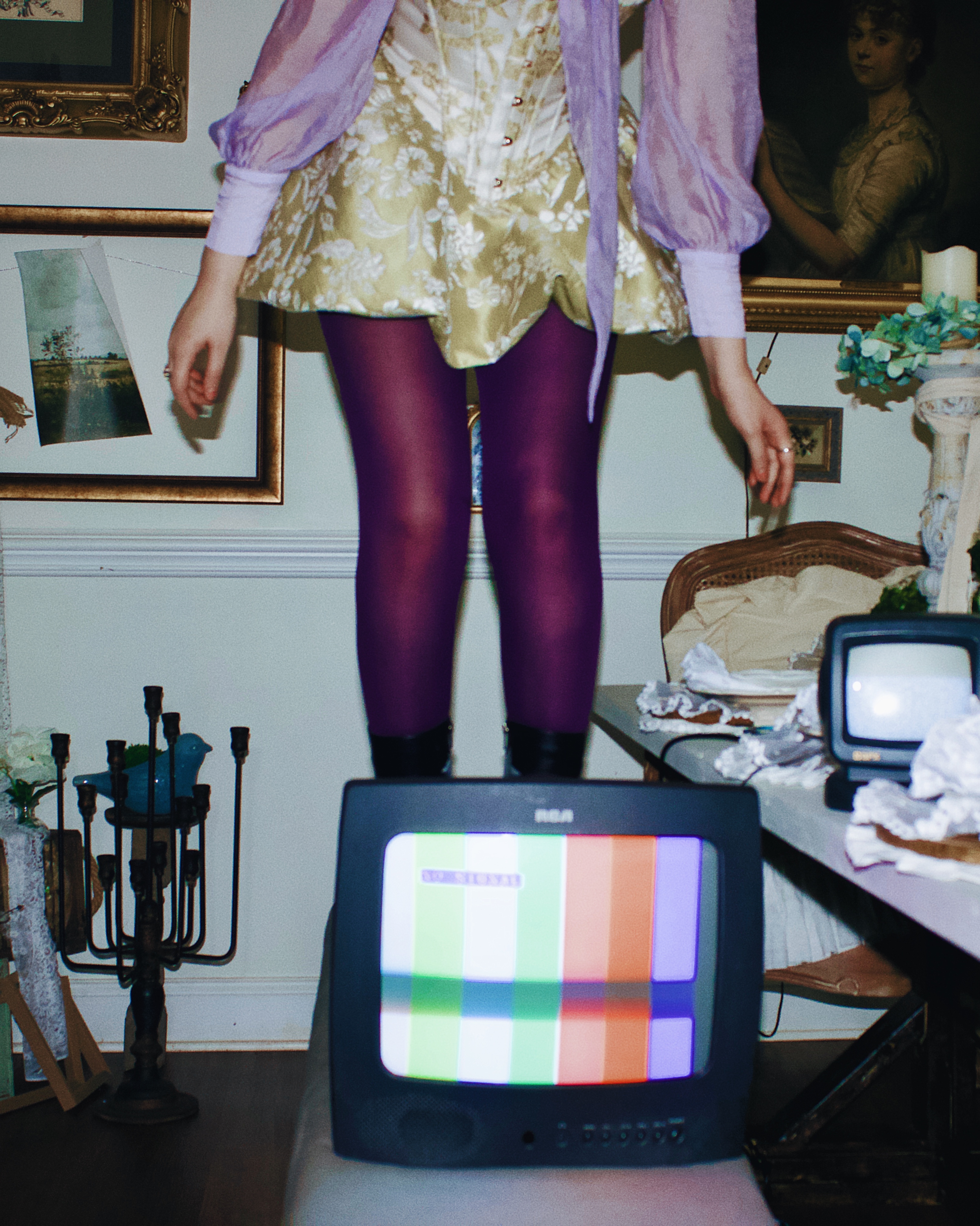 A person standing on a vintage television set displaying colored test patterns, dressed in a floral dress and purple tights, with various decorative items and art in the background.