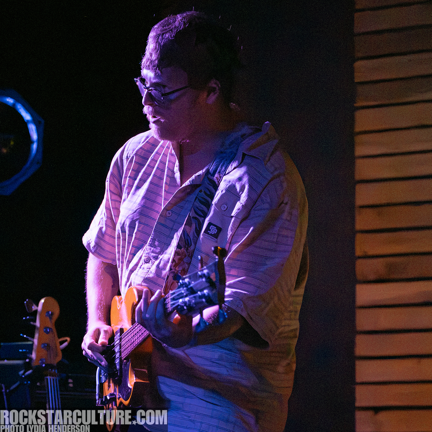 A musician playing an electric guitar on stage, wearing glasses and a striped shirt, with a focused expression.