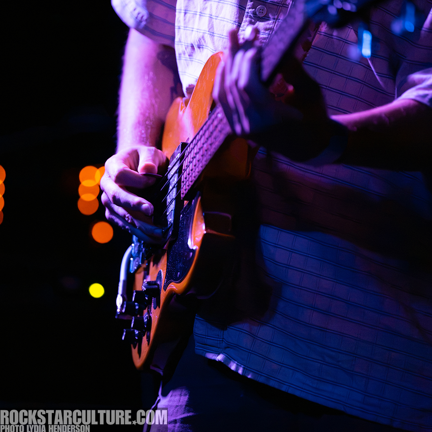 Close-up of a musician's hands playing an electric guitar on stage, with colorful lights in the background.