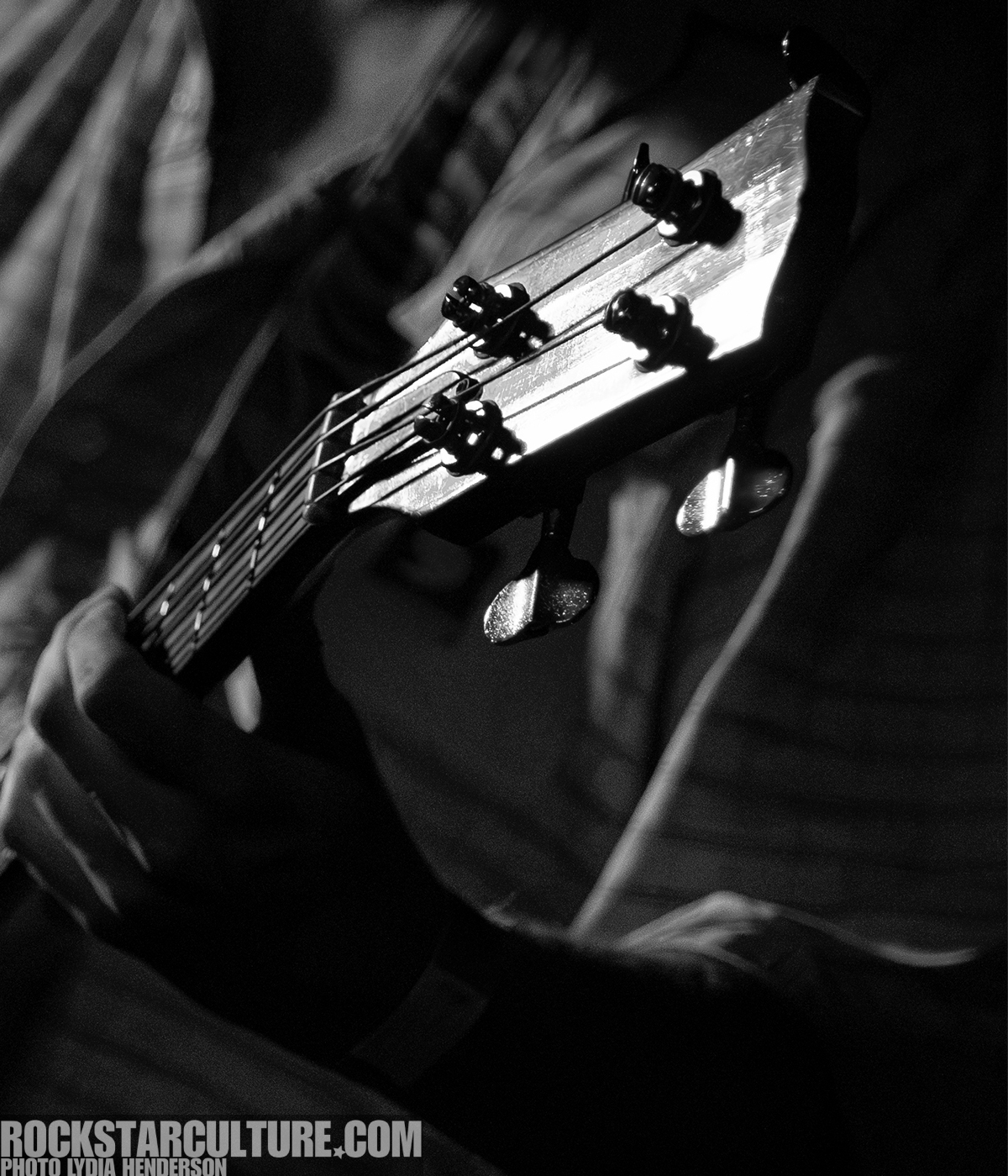 Close-up of a person's hand holding an acoustic guitar, focusing on the neck and tuning pegs in a dimly lit setting.