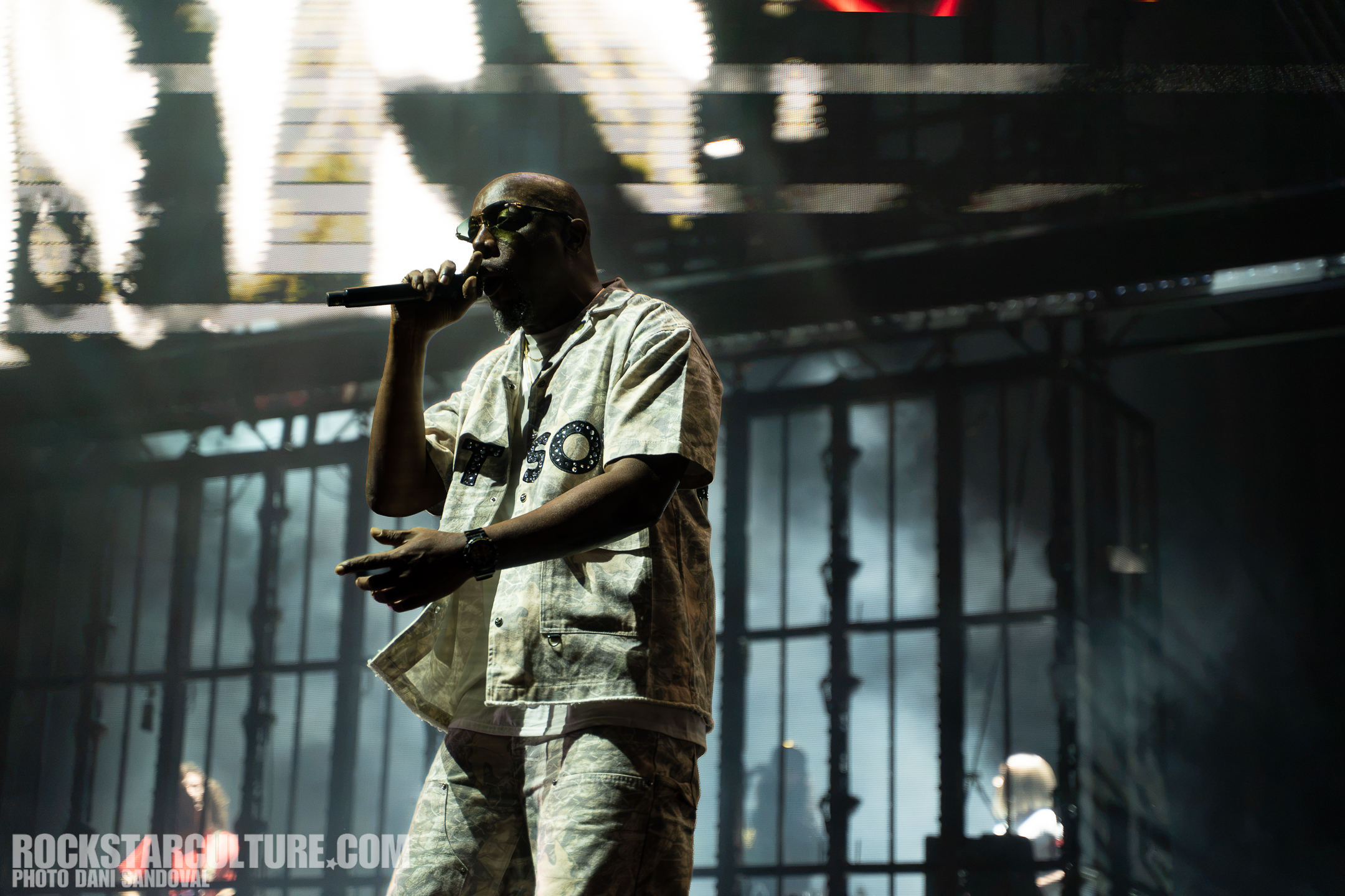 A performer singing into a microphone on stage, wearing a patterned outfit, illuminated by dramatic lighting with a blurred background featuring a band member.