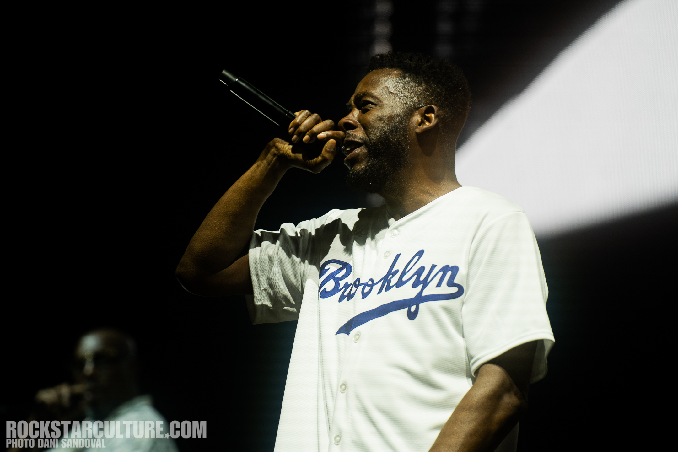 A performer passionately rapping into a microphone while wearing a white shirt with 'Brooklyn' printed on it, set against a dark background with stage lights.