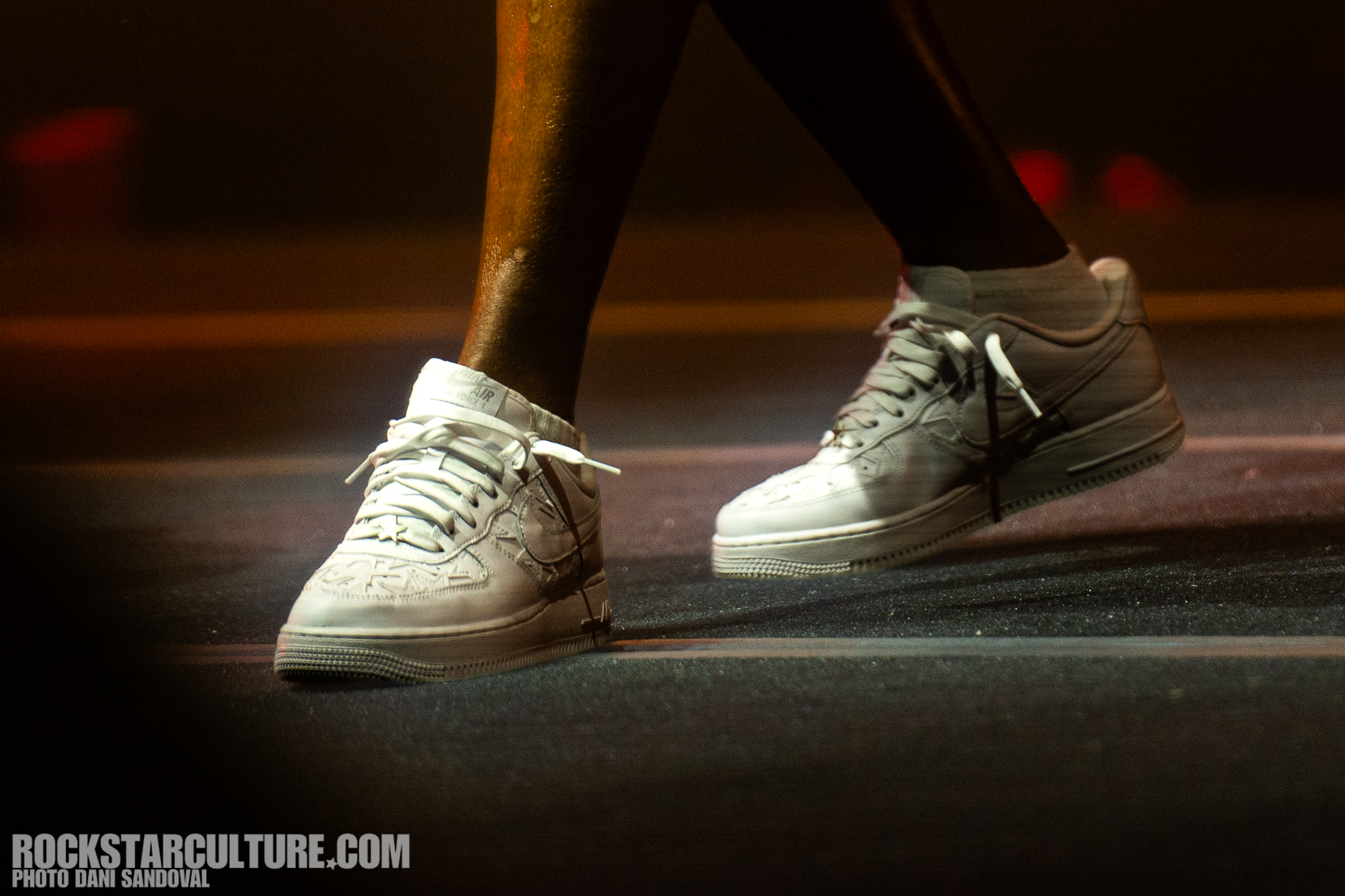 Close-up of a person wearing white Nike Air Force 1 sneakers, with a blurred background, on stage.