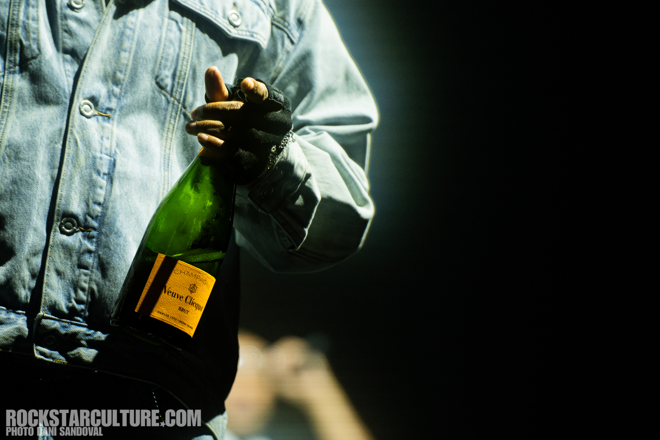 A person in a denim jacket holding a bottle of champagne against a dark background.