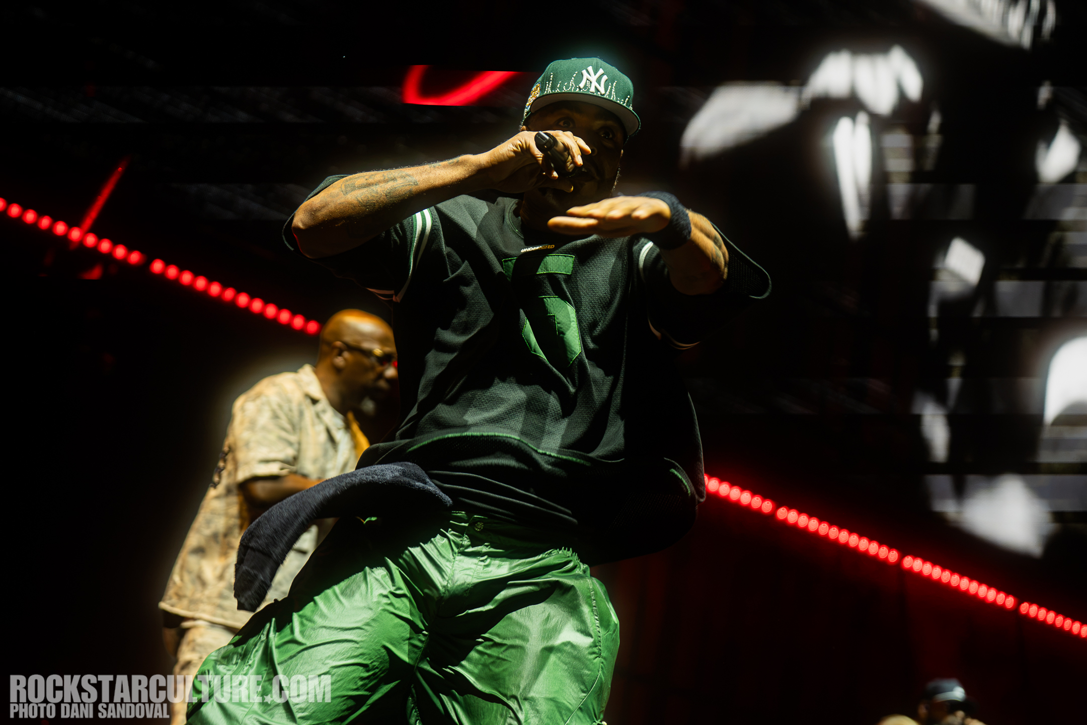 A performer in a black jersey and green pants rapping on stage, with a microphone in hand. A second performer is partially visible in the background, dressed in a light-colored shirt.
