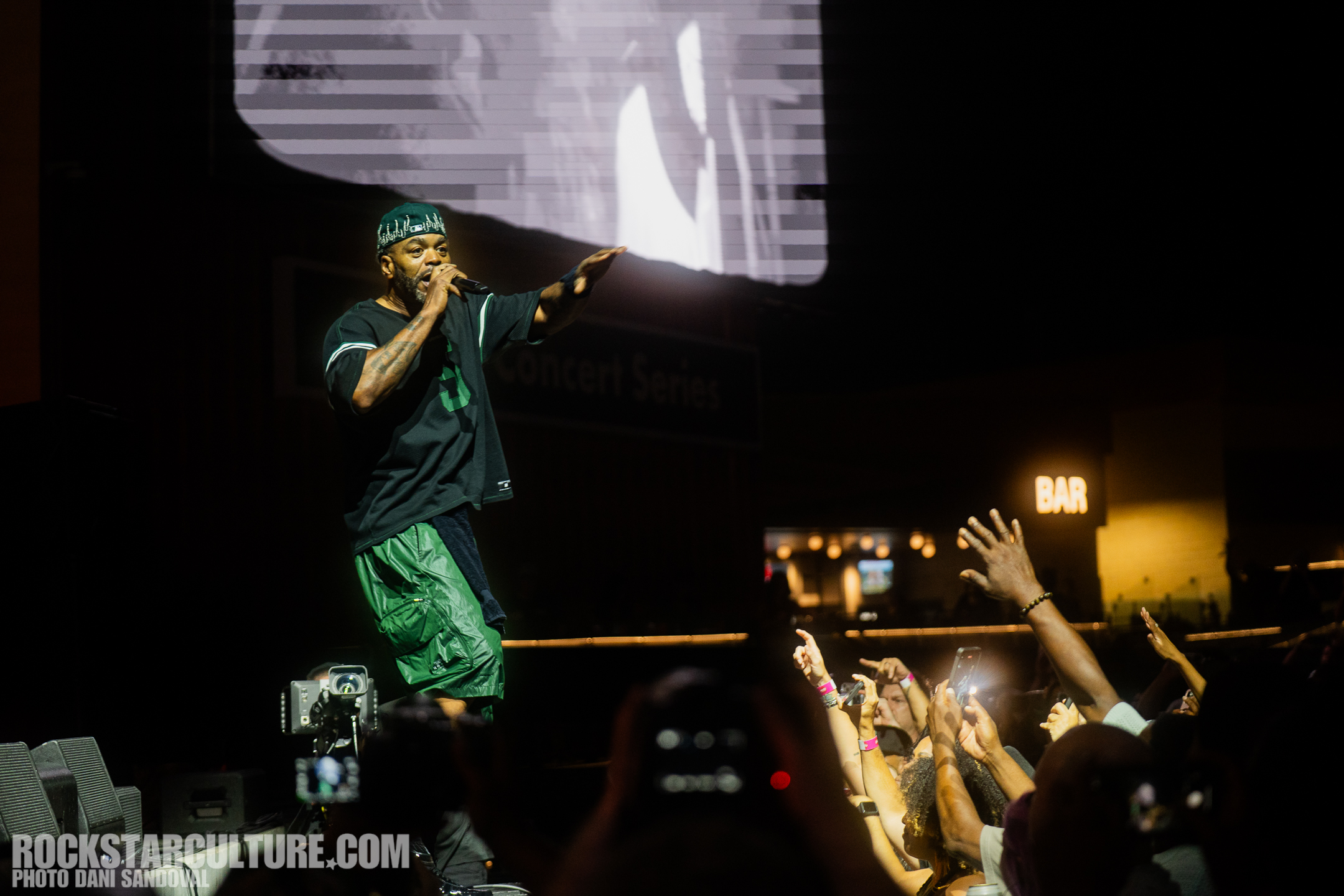 A performer on stage engaging with a lively crowd at a concert. The artist is wearing a black shirt and green shorts, and is holding a microphone while gesturing. The audience is reaching out with hands and recording the performance with their phones.